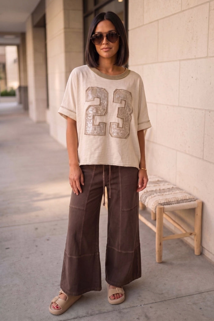 Neutral oversized boho boutique tee with sequin 23 graphic paired with olive wide leg lounge pants and slip-on sandals for a casual everyday street style look.