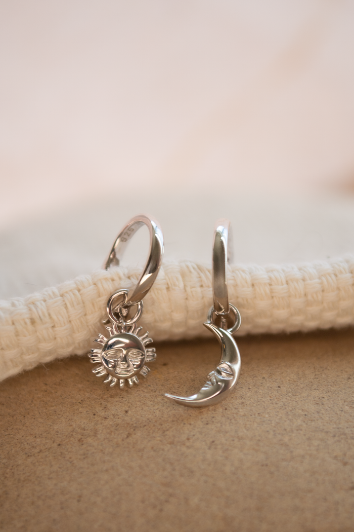 A pair of silver huggie hoop earrings with dangling sun and crescent moon charms, displayed on a textured beige surface.