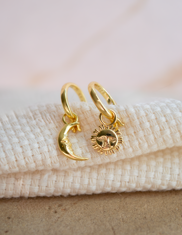A pair of gold-toned hoop earrings with dangling crescent moon and sun charms, displayed on a folded white textured fabric against a soft beige background.