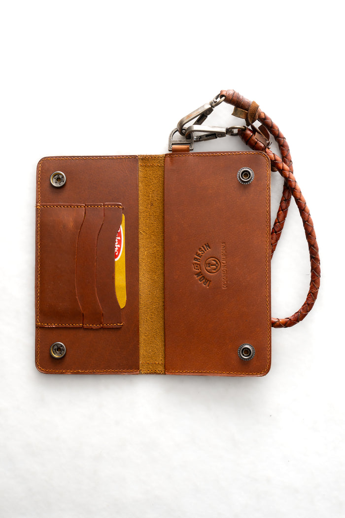Open brown leather wallet with card slots, snap buttons, and braided strap on white background. Card partially visible in slot.