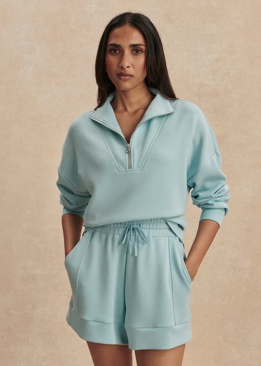 Woman wearing a blue 1/2 zip sweatshirt with matching shorts against a tan background