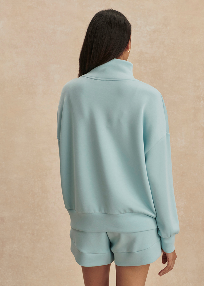 Woman turned around wearing a blue 1/2 zip sweatshirt with matching shorts against a tan background