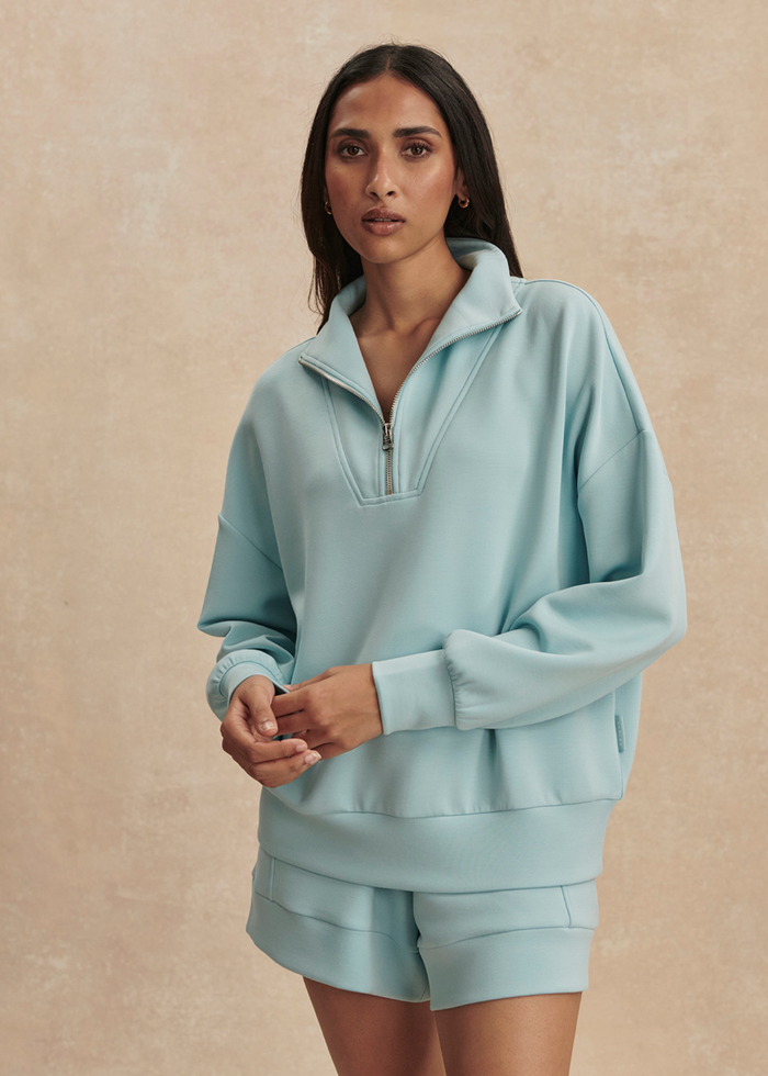Woman wearing a blue 1/2 zip sweatshirt with matching shorts against a tan background