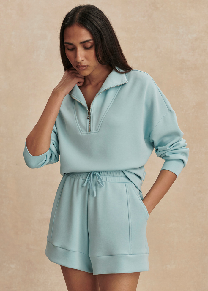 Woman wearing a blue 1/2 zip sweatshirt with matching shorts against a tan background